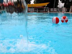 Das Freibad ist eröffnet. Wasserbälle schwimmen im Nichtschwimmerbecken vor einer Regendusche.