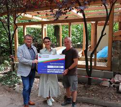 Ein großer symbolsicher Scheck wird von einer Frau und zwei Männern vor der Baustelle für ein neues Kleintierhaus festgehalten.