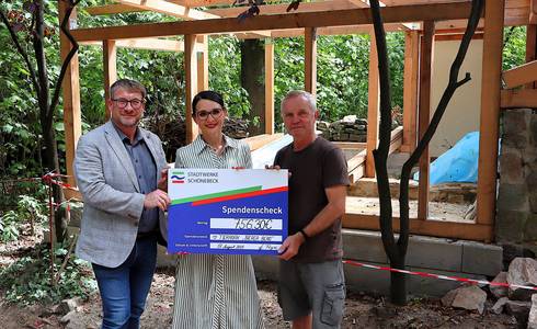 Ein großer symbolsicher Scheck wird von einer Frau und zwei Männern vor der Baustelle für ein neues Kleintierhaus festgehalten.